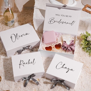 May include: White gift boxes with names Olivia, Rachel, and Chaya, and a box that says "Will you be my Bridesmaid?" with gifts inside. The boxes have gray ribbons. The open box contains heart-shaped sunglasses and a pink item.