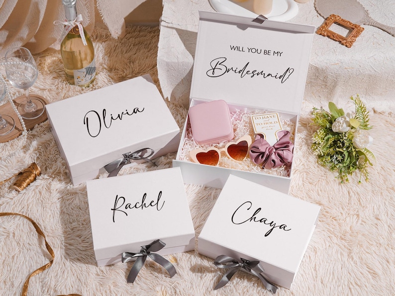 May include: White gift boxes with names Olivia, Rachel, and Chaya, and a box that says "Will you be my Bridesmaid?" with gifts inside. The boxes have gray ribbons. The open box contains heart-shaped sunglasses and a pink item.