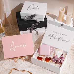 May include: Gift boxes in white, black, and pink, with names Cecilia and Amanda on them. One box is open, revealing a pink square, heart-shaped sunglasses, and a scrunchie. The open box reads "Will you be my Made of honor?"