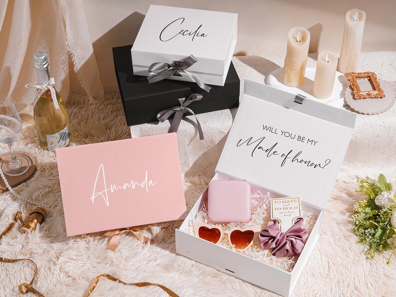 May include: Gift boxes in white, black, and pink, with names Cecilia and Amanda on them. One box is open, revealing a pink square, heart-shaped sunglasses, and a scrunchie. The open box reads "Will you be my Made of honor?"