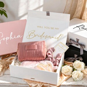 May include: Gift boxes with bridesmaid proposal theme. The boxes are white and pink, with gold and white text. Inside the boxes are a pink cosmetic bag, sleep mask, and scrunchies. White roses and decorative elements are also present.