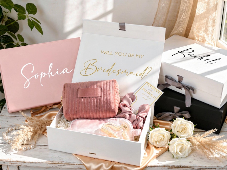 May include: Gift boxes with bridesmaid proposal theme. The boxes are white and pink, with gold and white text. Inside the boxes are a pink cosmetic bag, sleep mask, and scrunchies. White roses and decorative elements are also present.