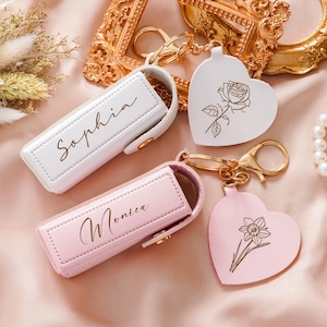 Può includere: Due custodie rettangolari per rossetto con chiusure a scatto dorate e portachiavi a forma di cuore. Una custodia è bianca con il nome Sophia e una rosa, l'altra è rosa chiaro con il nome Monica e l'illustrazione di un narciso.