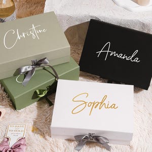 May include: Four gift boxes in shades of green, black, and white, each with a name in elegant script: "Christine," "Amanda," and "Sophia." The boxes are tied with satin ribbons. The boxes are on a fluffy, white surface.