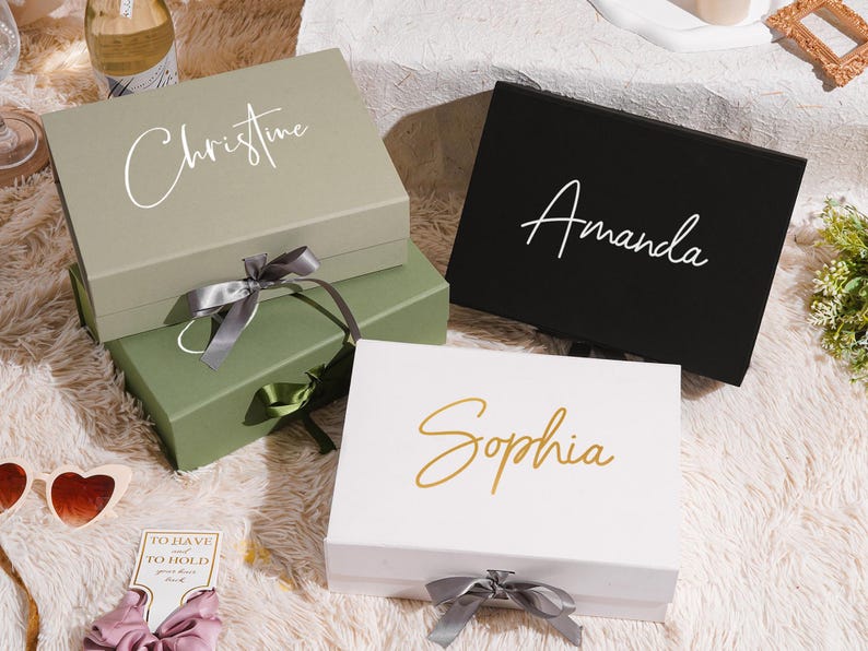 May include: Four gift boxes in shades of green, black, and white, each with a name in elegant script: "Christine," "Amanda," and "Sophia." The boxes are tied with satin ribbons. The boxes are on a fluffy, white surface.