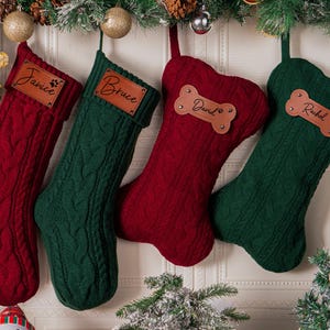 May include: Four cable-knit Christmas stockings in deep red and forest green. Two stockings are traditional boot-shaped with rectangular leather tags labeled Janice and Bruce. Two are bone-shaped with leather tags labeled David and Rachel.