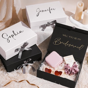 May include: Gift boxes, one black and two white, with names in cursive script. The black box is open, revealing gifts. The white boxes have gray ribbons. The open box has the text "Will you be my Bridesmaid?"