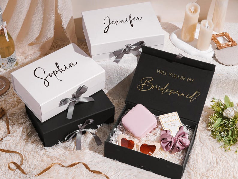 May include: Gift boxes, one black and two white, with names in cursive script. The black box is open, revealing gifts. The white boxes have gray ribbons. The open box has the text "Will you be my Bridesmaid?"