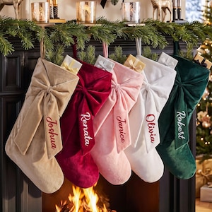 May include: Five velvet Christmas stockings in beige, deep red, light pink, white, and dark green, each featuring a large fabric bow and personalized embroidery with the names Joshua, Renata, Janice, Olivia, and Robert, hanging from a fireplace.