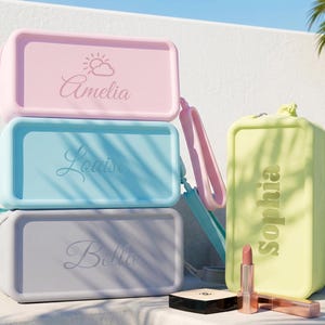 May include: A stack of three rectangular silicone pouches in pastel pink, light blue, and gray, each personalized with a name and a sun icon. Beside them stands a yellow pouch labeled Sophia, with a lipstick and compact case nearby.
