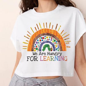May include: White crop top featuring a colorful rainbow design with a green caterpillar and the text "We Are Hungry FOR LEARNING". The design includes orange, brown, blue, and yellow colors. The shirt is a casual style.