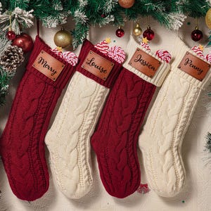 May include: Four knitted Christmas stockings in alternating deep red and cream colors, each featuring a thick cable knit pattern and a folded cuff. A rectangular brown leather patch is attached to each cuff, engraved with the names Merry, Louise, Jessica, and Penny.
