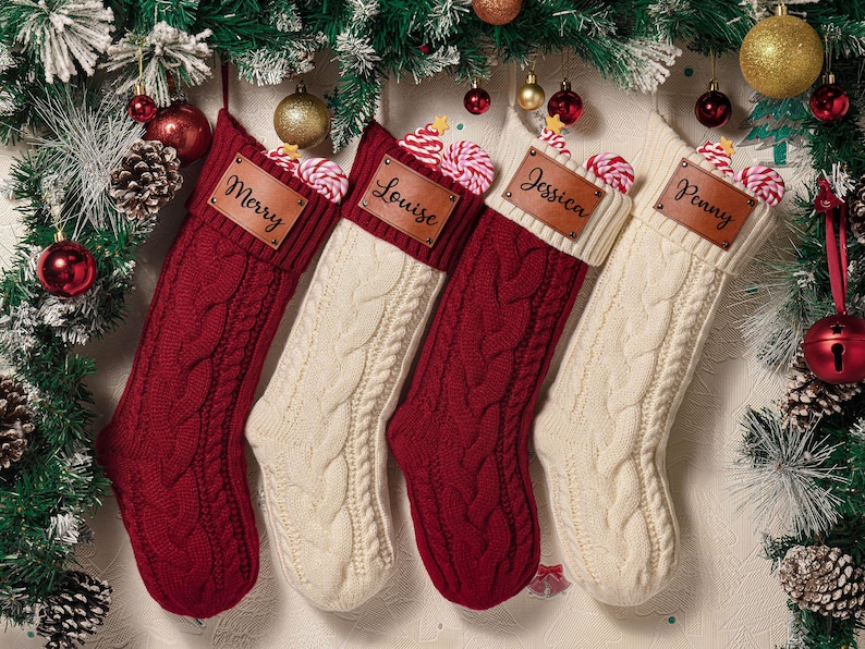 May include: Four knitted Christmas stockings in alternating deep red and cream colors, each featuring a thick cable knit pattern and a folded cuff. A rectangular brown leather patch is attached to each cuff, engraved with the names Merry, Louise, Jessica, and Penny.