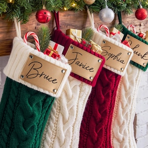Peut inclure: Quatre chaussettes de Noël en tricot torsadé vert, blanc et rouge, chacune avec un revers blanc et une étiquette rectangulaire en cuir beige fixée par quatre rivets argentés. Les étiquettes portent les noms Bruce, Janice, Sophia et Chunk.