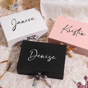 May include: Three gift boxes in white, pink, and black, each with a name in elegant script: "Janice", "Kristin", and "Denise". The boxes are tied with ribbons and sit on a fluffy, white surface. A perfect gift for a special occasion.