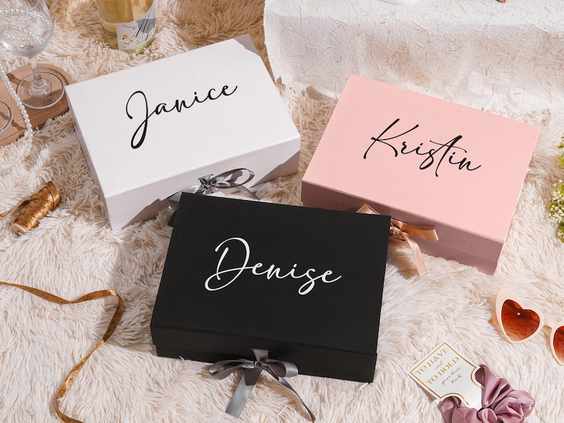 May include: Three gift boxes in white, pink, and black, each with a name in elegant script: "Janice", "Kristin", and "Denise". The boxes are tied with ribbons and sit on a fluffy, white surface. A perfect gift for a special occasion.