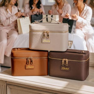 May include: Three rectangular vanity cases with top handles and gold-tone zippers. The top case is beige with the name Linda, the bottom left is tan with the name Janice, and the bottom right is dark brown with the name Rachel embossed in gold.