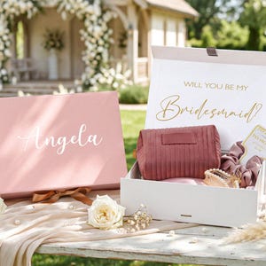 May include: A pink gift box with the name "Angela" in white script, next to an open box with a cosmetic bag, scrunchies, and a card that says "To Have and to Hold". The open box has the text "Will You Be My Bridesmaid?"