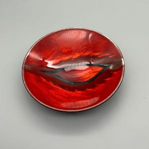 Può includere: Un piatto in ceramica rosso con un design nero. Il piatto ha una leggera curvatura ed è perfetto per servire spuntini o per contenere gioielli.