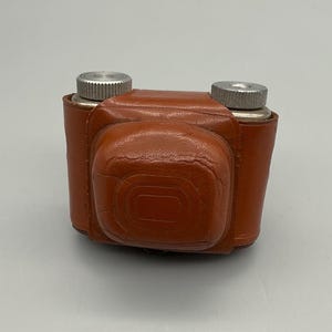Puede incluir: Una funda de cámara vintage de cuero marrón con dos diales plateados en la parte superior. La funda tiene forma rectangular con bordes redondeados y una superficie texturizada. La funda de la cámara está diseñada para proteger una cámara.