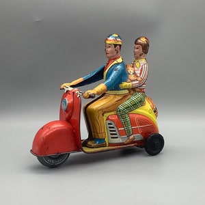 May include: A red, white, and blue toy scooter with two figures riding on it. The figures are wearing colorful clothing and one is holding a small toy animal.