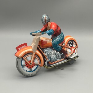 May include: A vintage toy motorcycle with a rider wearing a helmet and racing suit. The motorcycle is orange and red with a black and white tire. The motorcycle has the word "Technofix" printed on the side and the number "2" on the back wheel.