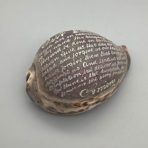 Peut inclure: Une coquille de mer avec une inscription manuscrite à l'encre argentée. L'inscription dit "Notre Père qui es aux cieux, que ton nom soit sanctifié. Que ton règne vienne. Que ta volonté soit faite sur la terre comme au ciel. Donne-nous aujourd'hui notre pain quotidien. Pardonne-nous nos offenses, comme nous pardonnons aussi à ceux qui nous ont offensés. Ne nous laisse pas succomber à la tentation, mais délivre-nous du mal. Car le royaume, la puissance et la gloire sont à toi, pour les siècles des siècles. Amen."