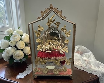 May include: An ornate display case featuring a red velvet cushion, embellished with gold-toned floral details and a bird motif. The glass-fronted frame is on a wooden surface, with a bouquet of white roses in the background.