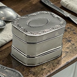 May include: A silver rectangular box with an ornate lid and a decorative border. The box has a layered design and sits on a wooden surface. Silverware is visible in the background.