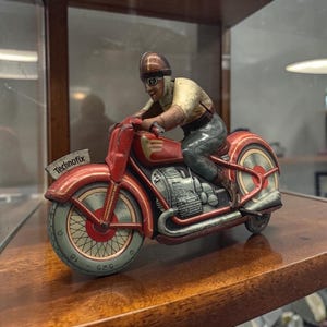 May include: Vintage red Technofix motorcycle toy with a rider wearing a helmet and goggles. The toy has a detailed engine and wheels with a cream and red color scheme. The Technofix logo is visible on the side.