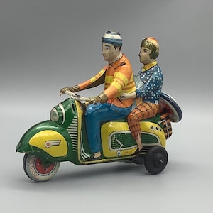 May include: A green and yellow toy scooter with a wind-up key. Two figures are riding the scooter, one wearing a blue and orange striped shirt and a blue and white hat, and the other wearing a blue and white polka dot shirt and a red and white hat. The scooter has the word "TECHNOMATIC" printed on the side.