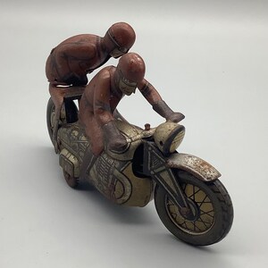 Op de afbeelding: Een vintage tinnen speelgoedmotorfiets met twee rijders. De motorfiets is geschilderd in bruin, zilver en zwart met een gedetailleerd ontwerp. De rijders dragen bruine outfits en helmen.