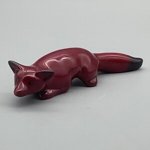 Puede incluir: Una pequeña figura de zorro de cerámica roja con una punta negra en la cola. El zorro está acostado con la cabeza girada hacia un lado.