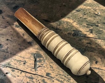 May include: A handcrafted, light-colored scoop with a cylindrical handle. The scoop's bowl is a lighter shade of brown, while the handle features dark brown bands and a rounded base. The item is made of wood and is resting on a wooden surface.