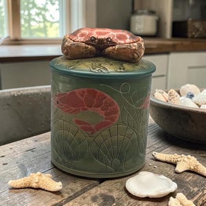 May include: A ceramic jar with a crab lid. The jar is green with a red shrimp design and shell patterns. The crab lid is brown and red. The jar is on a wooden surface with seashells and starfish.