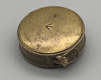 May include: A brass measuring tape with a white fabric ribbon and a small arrow on the top. The tape is closed and the ribbon is visible.