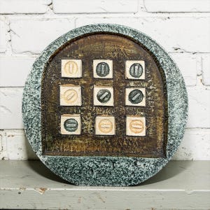 May include: A circular wall plaque with a textured, mottled green and gray border surrounding a dark brown, weathered central panel. Nine square tiles are arranged in a three-by-three grid, each featuring a raised diagonal bar in shades of cream, tan, or dark green.