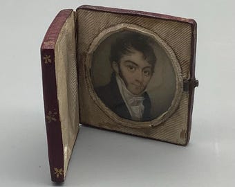 May include: A small, red leather-bound book with a gold design on the cover. The book is open to reveal a portrait of a man in a white shirt and dark jacket. The portrait is framed in a circular, cream-colored frame.