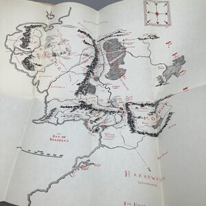 Puede incluir: Un mapa detallado de una tierra ficticia, con intrincadas ilustraciones de monta&ntilde;as, r&iacute;os y asentamientos. El mapa es principalmente en blanco y negro, con acentos rojos para nombres de lugares y caracter&iacute;sticas clave.