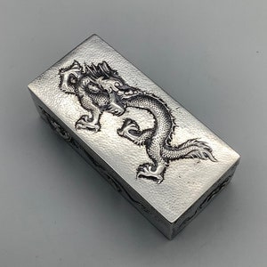 May include: A silver rectangular box with a raised, embossed design of a dragon on the top. The box has a textured surface.