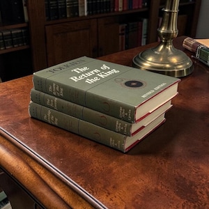 Puede incluir: Tres libros de tapa dura apilados sobre una superficie de madera. El libro superior se titula "The Return of the King" de J.R.R. Tolkien, con una cubierta verde y texto blanco. Los otros dos libros tambi&eacute;n son novelas de Tolkien. Los libros tienen bordes rojos.