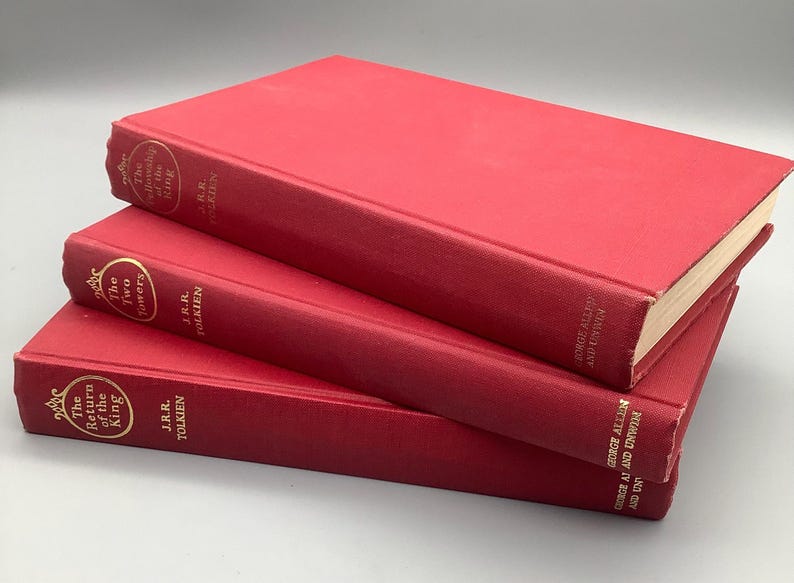 Puede incluir: Una pila de tres libros de tapa dura rojos. Los t&iacute;tulos son "La Comunidad del Anillo", "Las Dos Torres" y "El Retorno del Rey". Todos los libros son de J.R.R. Tolkien, con t&iacute;tulos dorados en los lomos.