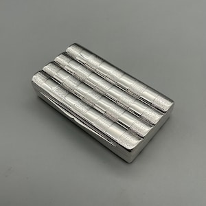 May include: A silver cigarette case with a geometric pattern. The case is rectangular and has a hinged lid.