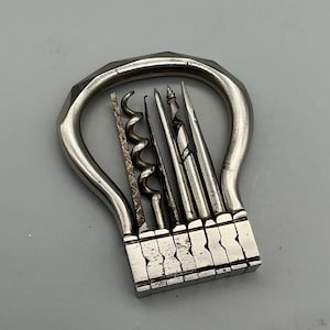 Pode incluir: Uma ferramenta multiuso vintage prateada com vários implementos, incluindo saca-rolhas e ferramentas pontiagudas. A ferramenta tem uma alça distinta e uma base retangular com várias ranhuras. O design geral sugere um foco na funcionalidade e no artesanato.