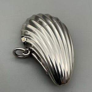May include: A silver-colored, clam-shaped container with a hinged lid and a small ring attached. The container has a ribbed design and a small gold-colored accent near the hinge. The overall shape is elongated and oval.