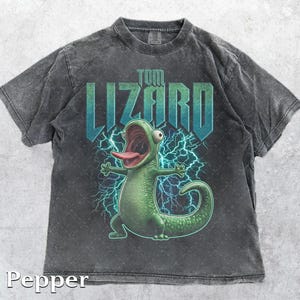 May include: A dark gray t-shirt featuring a cartoon lizard with its mouth open and tongue sticking out. The lizard is green with a blue lightning bolt background. The text "TOM LIZARD" is above the lizard in a blue and green font.