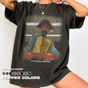 May include: A charcoal gray short-sleeve t-shirt featuring a graphic of a pirate character in a yellow vest and blue sleeves. The text reads Cap'n Clark's OTTOMAN EMPIRE with a ship wheel icon. Below, a color palette shows available pepper colors.