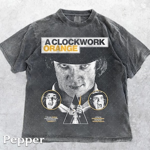 Puede incluir: Camiseta color pimienta con un gráfico de la película "La Naranja Mecánica". El diseño incluye el título de la película, un retrato y otras imágenes. La camiseta está hecha de un material suave.
