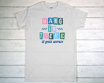 May include: White t-shirt with the text "HANG IN THERE it gets worse" in colorful block letters. The shirt has a crew neck and short sleeves. The text is in pink, blue, green, and teal.