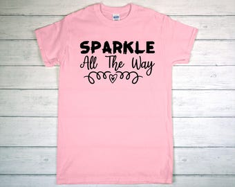 May include: A light pink t-shirt with the words "SPARKLE All The Way" in black lettering. The design includes a heart and decorative swirls. The shirt is made of a soft material.